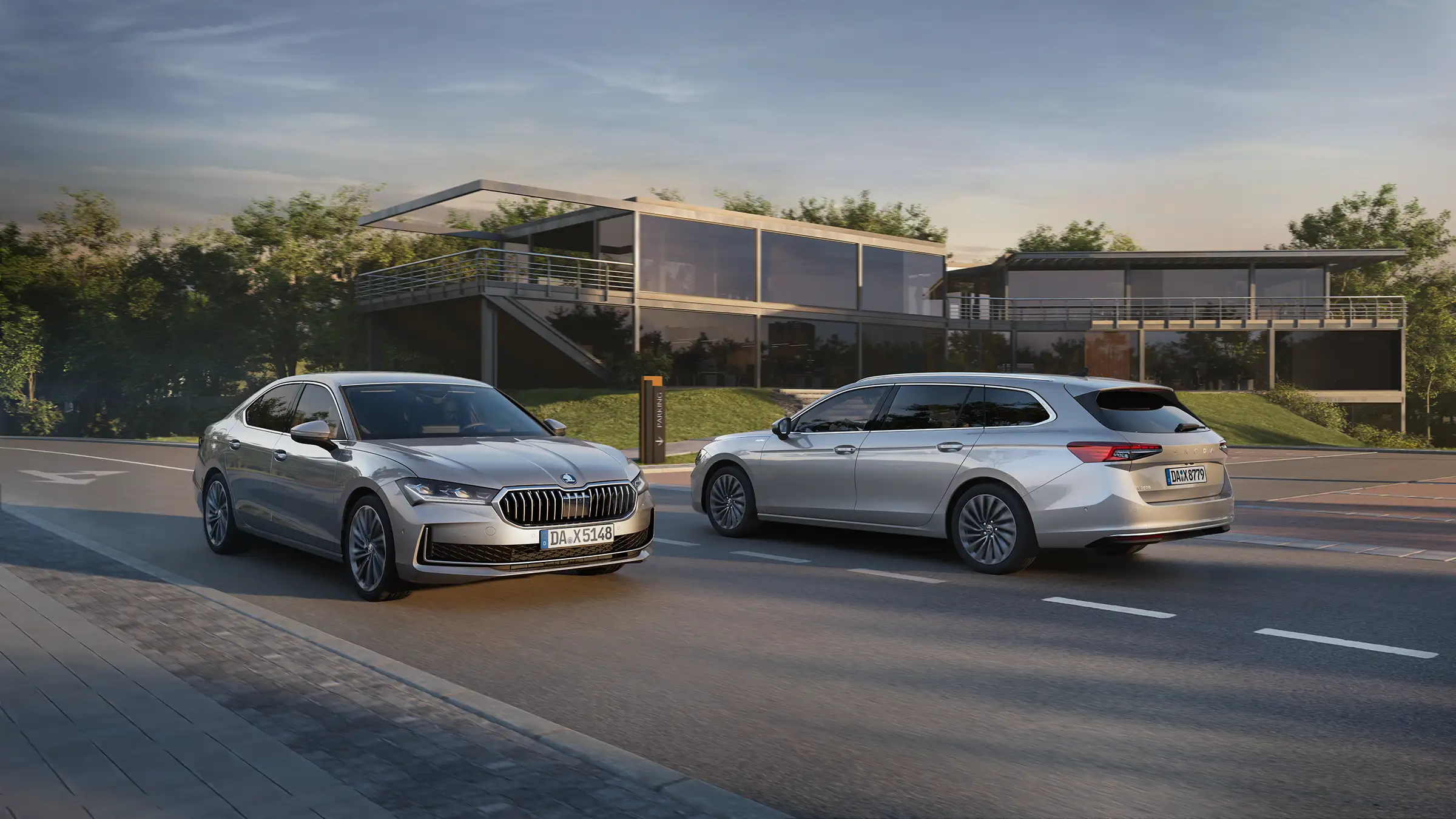 Zwei graue Autos fahren auf einer Straße vor modernen Gebäuden mit großen Glasfenstern. Die Umgebung ist grün und die Straße ist zweispurig mit klaren Fahrbahnmarkierungen.
