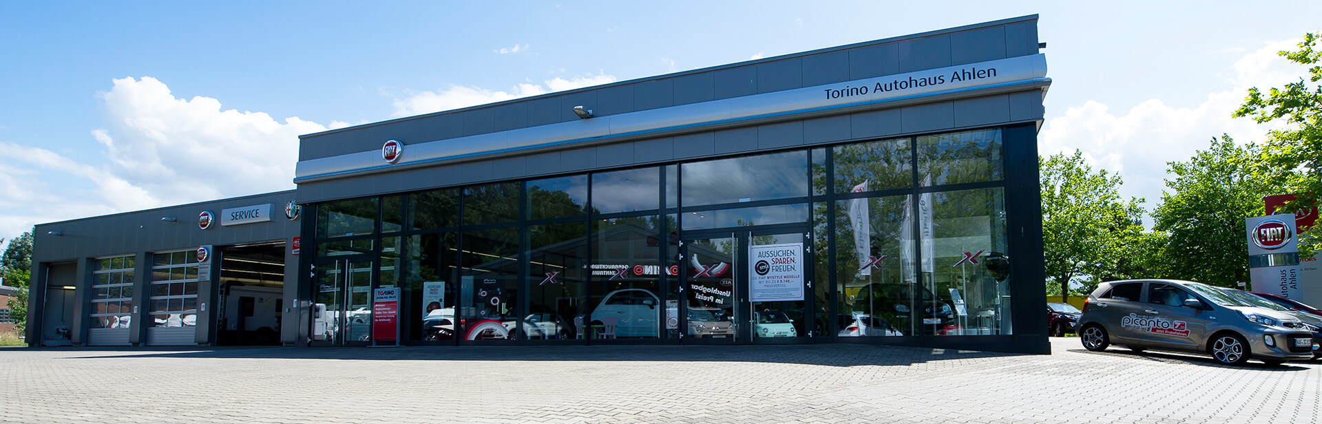 Außenansicht eines Autohauses mit großen Glasfenstern. Auf dem Gebäude steht „Torino Autohaus Ahlen“. Links ist der Service-Bereich mit Rolltoren zu sehen. Rechts vor dem Gebäude steht ein silbernes Auto. Im Hintergrund sind Bäume und ein Schild mit dem Fiat-Logo sichtbar. Der Himmel ist leicht bew
