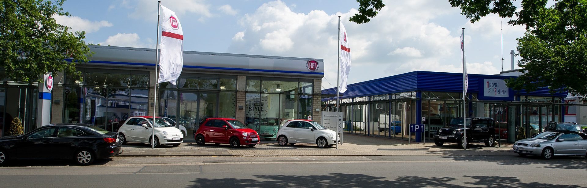Eine Autowerkstatt mit dem FIAT-Logo und mehreren geparkten Autos davor. Vor dem Gebäude wehen zwei weiße Flaggen mit dem FIAT-Logo im Wind. Auf dem Gehweg sind mehrere Autos zu sehen, darunter weiße und rote Fahrzeuge. Rechts sind weitere Autogebäude mit blauen Dächern erkenn