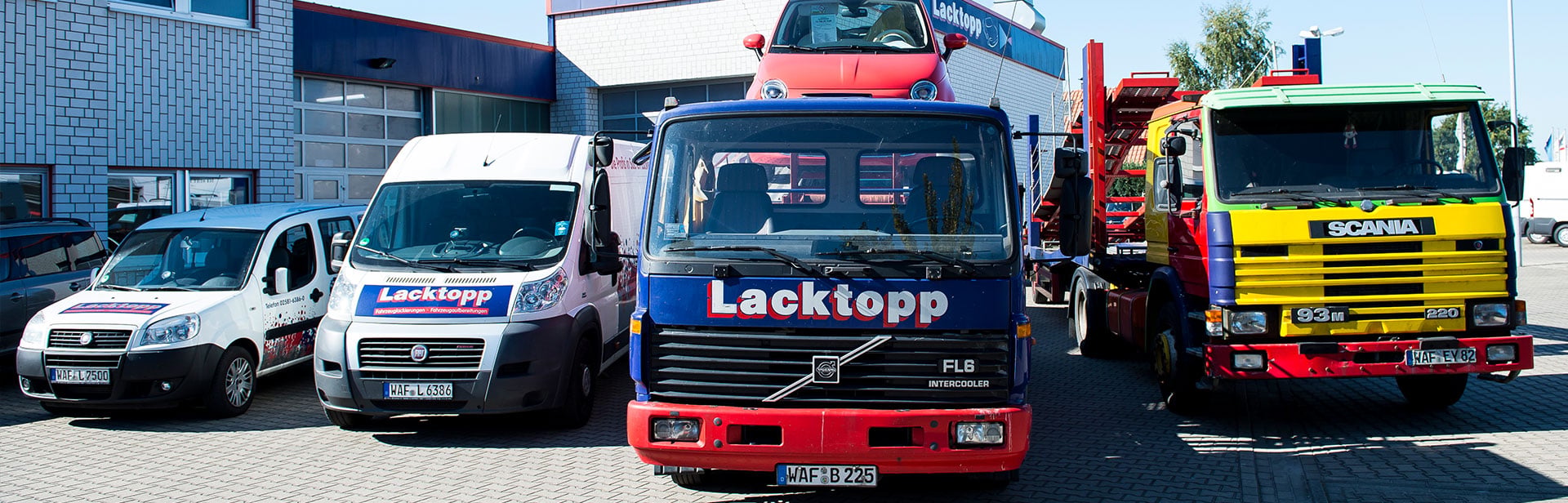 Eine Flotte von Fahrzeugen, die auf einem Parkplatz vor einem Gebäude geparkt sind. Links steht ein kleiner weißer Lieferwagen, daneben ein größerer weißer Transporter, beide mit Lacktopp-Beschriftung. Rechts daneben steht ein blauer Lastwagen mit Lacktopp-Schriftzug und weiter rechts ein b