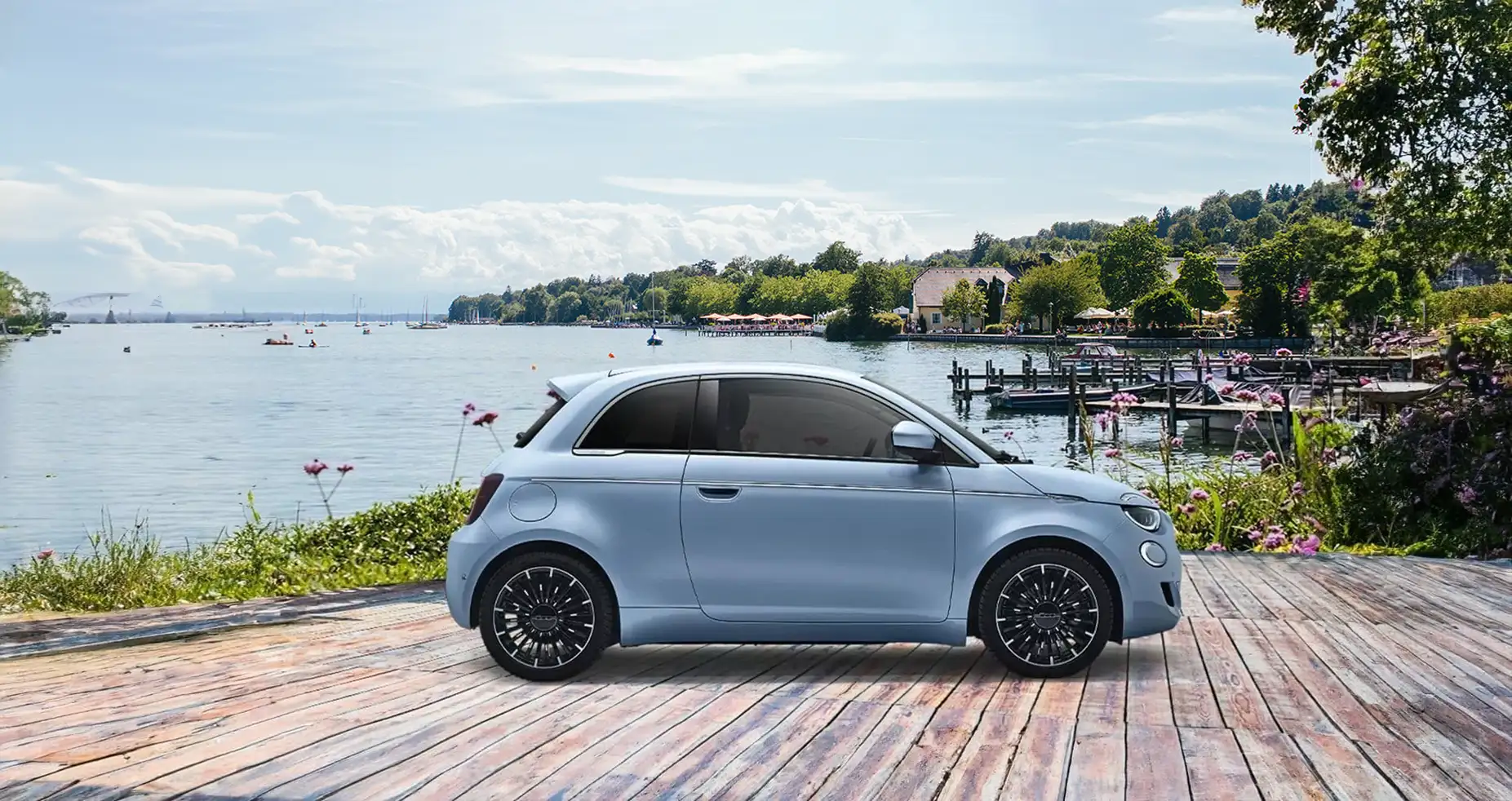 Ein hellblaues Auto steht auf einem Holzsteg vor einem malerischen See. Im Hintergrund sind Boote auf dem Wasser und eine Küstenlinie mit üppigen Bäumen und einem Gebäude mit Terrasse sichtbar. Der Himmel ist leicht bewölkt.
