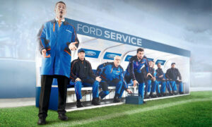 Ein Bild zeigt eine Gruppe von Männern in blauen Ford-Arbeitsanzügen, die in einer Wartezone mit der Aufschrift Ford Service sitzen. Einer von ihnen steht und spricht, während die anderen auf einer Bank sitzen oder sich vorbeugen. Der Hintergrund weist auf ein sportliches Umfeld hin, möglicherweise ein Stadion.