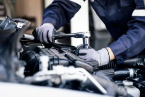 Eine Person in einem blauen Overall und grauen Handschuhen benutzt einen Schraubenschlüssel, um an einem Autoteil im Motorraum zu arbeiten.