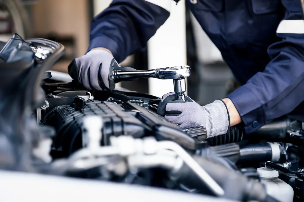 Eine Person in einem blauen Overall und grauen Handschuhen benutzt einen Schraubenschlüssel, um an einem Autoteil im Motorraum zu arbeiten.